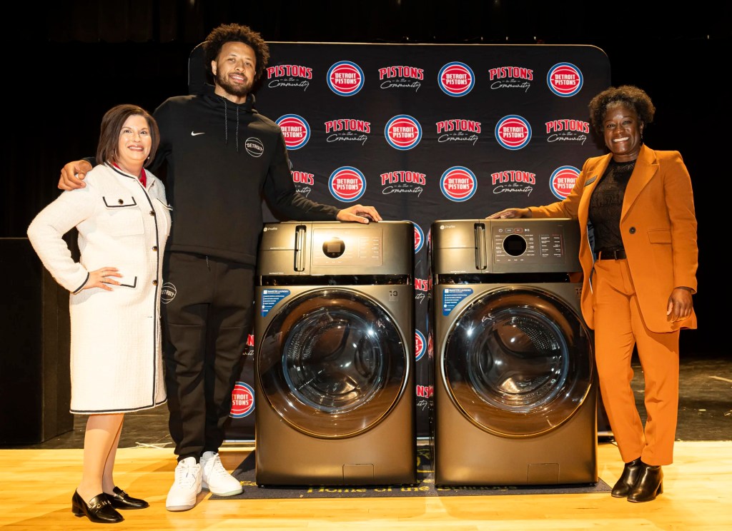 Two women from GE Appliances stand with Detroit Piston's basketball player Cade Cunningham in front of a GE Profile front load washer and dryer. A Detroit Pistons banner in partnership with Cade Cunningham are visible in the background. 